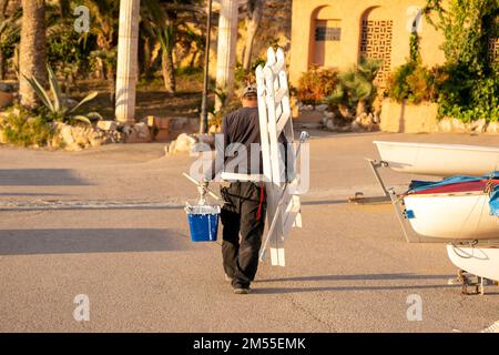 Vista posteriore di un dipendente anonimo che trasporta una scala e un secchio di vernice bianca che cammina sulla sabbia vicino alle barche durante il lavoro in spiaggia al mattino Foto Stock