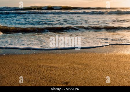 Onde di mare schiumose che si ondolano sulla spiaggia di sabbia illuminata al sole la mattina presto nel resort Foto Stock