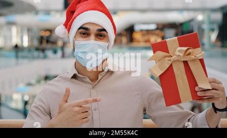 Uomo in Natale X-mas cappello ragazzo maschio in maschera di fronte Covid in piedi nel centro commerciale centro commerciale guardando la macchina fotografica che mostra regalo scatola di regalo avvolto in rosso Foto Stock