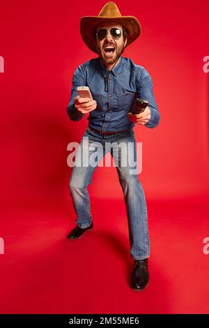 Giovane uomo eccitato in stile cowboy vestito divertirsi, posa isolato su sfondo rosso. Modello in cappello da cowboy e abiti in denim. Moda, emozioni Foto Stock