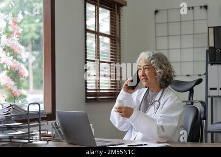 Medico asiatico senior femminile che utilizza il telefono cellulare parlando mentre si siede in ospedale ufficio. donna felice operatrice medica che ha una conversazione gioiosa con Foto Stock