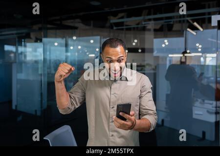 L'uomo d'affari è felice di celebrare la vittoria e il successo, l'uomo ispanico all'interno dell'ufficio tiene uno smartphone nelle sue mani ha ricevuto un messaggio gioioso notizia di trionfo tiene la mano in su. Foto Stock