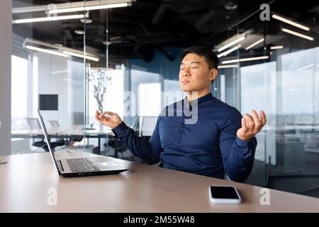 Stanco giovane uomo d'affari asiatico meditare sul lavoro. Si siede al tavolo in ufficio, si è preso una pausa, ha chiuso gli occhi, le mani nella posizione del loto, si rilassa. Foto Stock