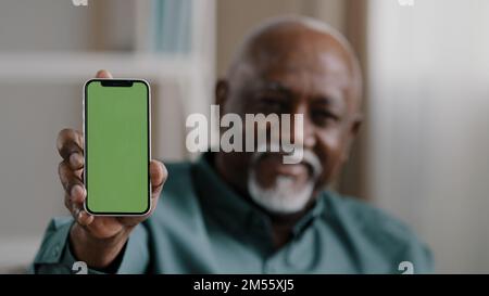 Vecchio uomo afro-americano biracial a casa seduto sul divano uomo d'affari in ufficio mostrando smartphone con schermo verde vuoto chromakey tenere Foto Stock