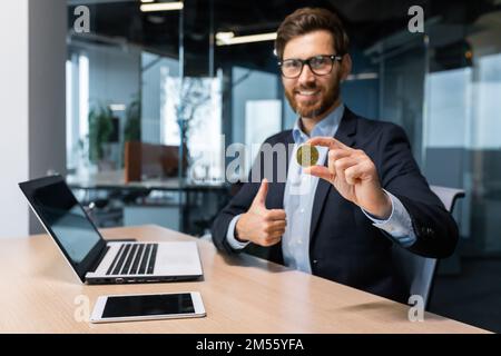 Ritratto di successo maturo imprenditore investitore, l'uomo mostra criptocurrency moneta d'oro per fotocamera e pollici fino saldamente, l'uomo lavora in borsa all'interno di ufficio online utilizza il computer portatile. Foto Stock