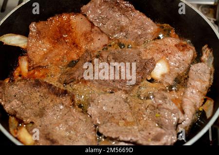 Pezzi di bistecca di carne alla griglia con olio, fette di cipolla, sale e pepe nero in una padella alla griglia, bistecche di manzo selezionate in fase di cottura, ricetta di carne d Foto Stock