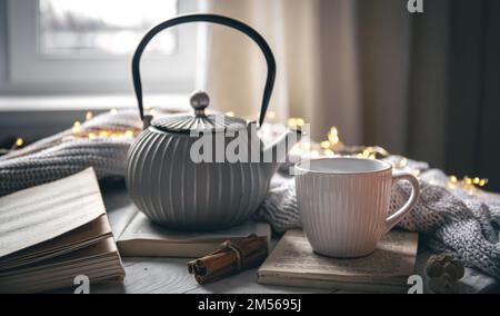 Composizione accogliente con teiera, tazza e libro su sfondo sfocato. Foto Stock