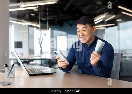 Felice giovane uomo asiatico seduto in ufficio con denaro contante e telefono. Gioire per la vittoria, lo stipendio, piazzare scommesse online, festeggiare. Foto Stock
