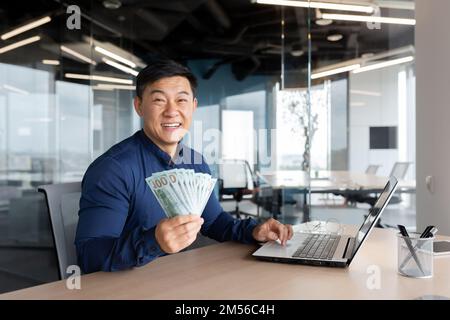 Felice giovane uomo d'affari asiatico seduto in ufficio al tavolo tenendo denaro contante nelle sue mani, mostrando alla fotocamera, sorridendo. Funziona su un computer portatile e documenti. Foto Stock