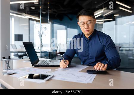 Ritratto di un serio giovane ragioniere asiatico, banchiere. Lavora in ufficio con documenti, computer portatile, calcolatrice. Foto Stock