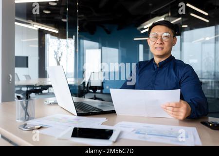 Ritratto di un giovane architetto, designer, ingegnere asiatico. Seduto alla scrivania in ufficio, tenendo i documenti. Lavora con documenti, progetti, piani. Guarda la telecamera, sorride. Foto Stock