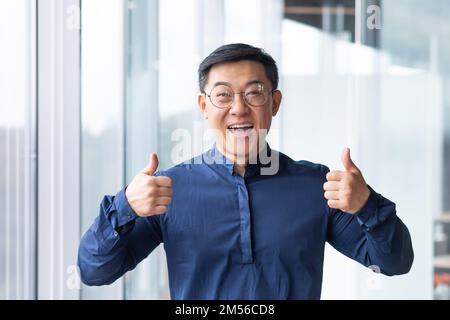 Ritratto di un giovane felice e bello uomo asiatico, uomo d'affari, studente in occhiali e una camicia blu in piedi in ufficio, guardando la macchina fotografica, sorridendo, mostrando le sue mani super. Foto Stock