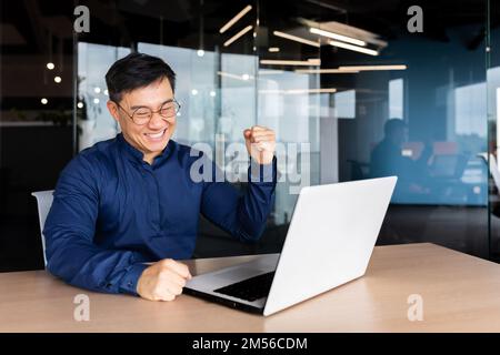 Un giovane asiatico lavora in ufficio su un portatile online. È felice, guarda il monitor, fa un gesto di sì con la mano. Foto Stock
