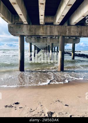 Un colpo verticale di onde che colpiscono la riva di una spiaggia vista da sotto un molo di legno Foto Stock