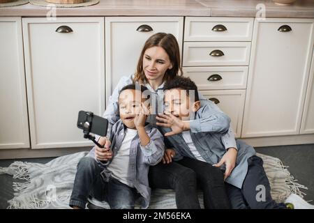 Felice famiglia multinazionale di madre e due figli seduti sul pavimento caldo in cucina leggera durante il fine settimana, riposarsi e scattare foto positive con selfie stick e le mani ondulate. Foto Stock