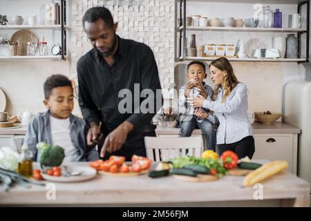 Fuoco selettivo sull'amore del bambino e del bambino del bambino del bambino del bambino del bambino piccolo che si diverte insieme nella cucina. Vista sfocata del padre africano che insegna a un altro piccolo figlio prescolare a tritare le verdure preparare l'insalata per il pranzo Foto Stock