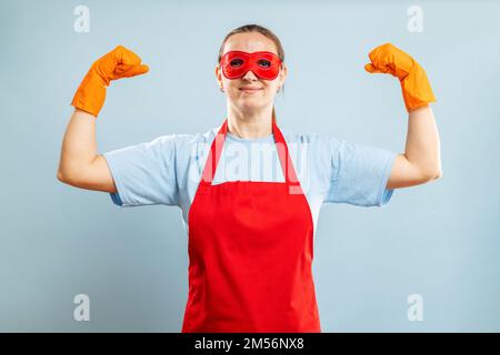 Giovane casalinga fiduciosa nella pulizia del costume supereroe su sfondo blu Foto Stock