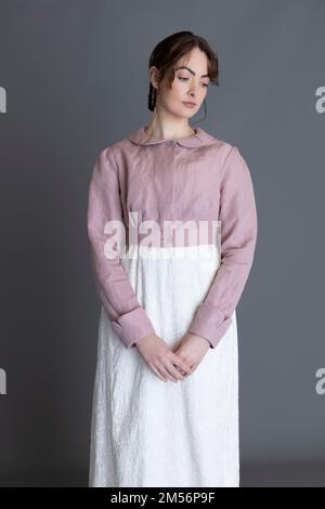 Una donna del periodo Regency che indossa un abito ricamato in crema con spencer in lino rosa sullo sfondo di un semplice studio Foto Stock