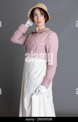 Una donna del periodo Regency che indossa un abito ricamato in crema con spencer in lino rosa sullo sfondo di un semplice studio Foto Stock