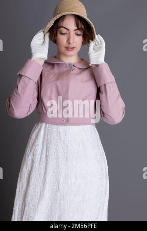 Una donna del periodo Regency che indossa un abito ricamato in crema con spencer in lino rosa sullo sfondo di un semplice studio Foto Stock