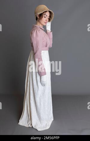 Una donna del periodo Regency che indossa un abito ricamato in crema con spencer in lino rosa sullo sfondo di un semplice studio Foto Stock