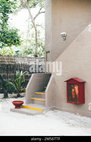 Ingresso casa di design tropicale con sabbia sul pavimento, recinzione di canna del residence, piante, uno strumento antincendio, una ciotola per il lavaggio dei piedi, linee gialle Foto Stock