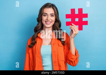 Ritratto fotografico di giovane donna attraente in possesso di hashtag icona influencer indossare abito arancione alla moda isolato su sfondo di colore blu Foto Stock