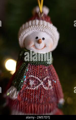 Un ornamento raffigurante un pupazzo di neve vestito di maglione rosso stagionale e sciarpa verde pende da un albero di Natale illuminato. Foto Stock