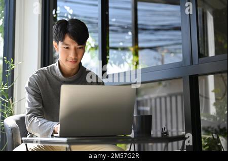 Giovane uomo d'affari asiatico intelligente e professionale che lavora in remoto presso la caffetteria, concentrandosi sul suo lavoro sul computer portatile. Foto Stock
