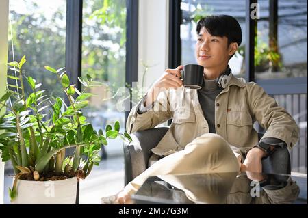Maschio asiatico di successo e bello in abiti casual ama avere il caffè del mattino al bar, guardando lontano dalla macchina fotografica, sognando ad occhi aperti il suo Foto Stock