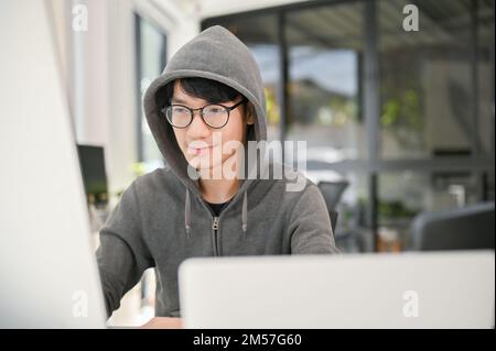 Smart e nerd giovane asiatico maschio sviluppatore o programmatore sito web indossando occhiali e felpa con cappuccio che si concentra sul suo compito sul computer, lavorando nel Foto Stock