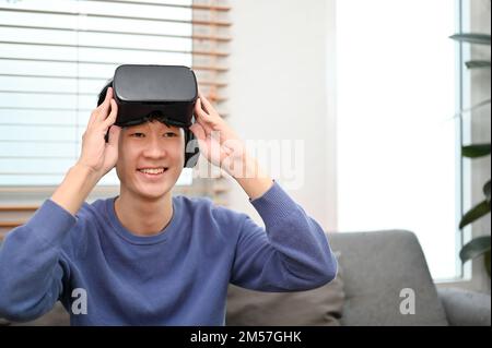 Giovane uomo asiatico gioioso che indossa occhiali VR e gioca a videogame VR nel suo salotto. Gadget per la realtà virtuale e concetto di stile di vita urbano Foto Stock