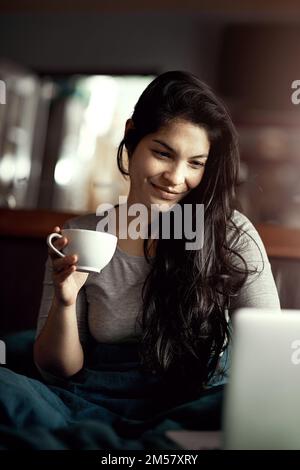 Non si stanca mai di guardare le immagini del gatto. una giovane donna sorridente che usa il computer portatile mentre beve il tè a letto. Foto Stock