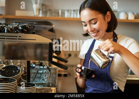 Primo piano di carina barista asiatica ragazza fare cappuccino, fare latte art in tazza con latte al vapore, in piedi in caffetteria dietro il bancone Foto Stock