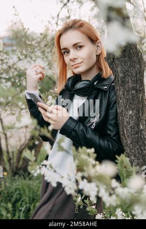 Una ragazza adolescente dai capelli rossi ascolta la musica in cuffie, configurate tramite telefono. Il tempo della fioritura primaverile di ciliegi. Foto Stock