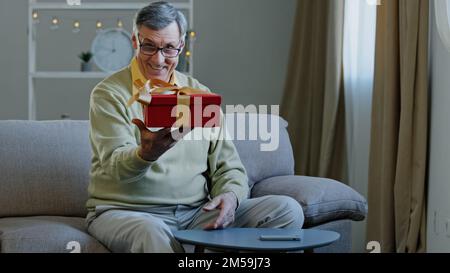 Buon vecchio caucasico nonno pensionato imballaggio rosso presente scatola legatura nastro. Grandad preparare il regalo al compleanno x-mas celebrazione seduta dentro Foto Stock