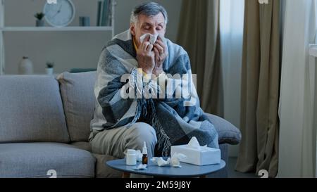 L'uomo anziano malato coperto con coperta calda soffre di starnuti naso che cola nel tovagliolo malsano vecchio nonno sensazione di febbre cattiva virus sintomi di malattia anziano Foto Stock