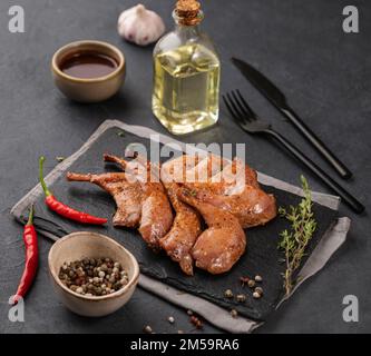 Cosce di quaglia grigliate alla griglia in marinata e spezie su ardesia nera con erbe fresche, olio e salsa su sfondo scuro. Alimentazione sana. Foto Stock