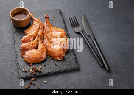 Prelibatezze. Cosce di quaglia grigliate alla griglia in marinata e spezie su ardesia nera con salsa su fondo scuro. Alimentazione sana. Spazio di copia. Foto Stock