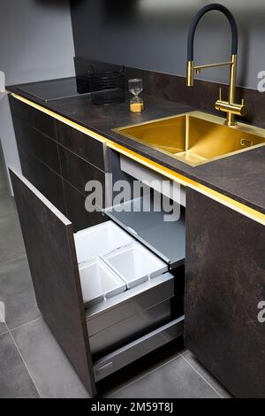 Armadio del cestino della cucina. Estrarre il cassetto della cucina per il cestino raccolta differenziata sotto il lavabo dorato e il rubinetto piano cottura elettrico in vetro nero Foto Stock