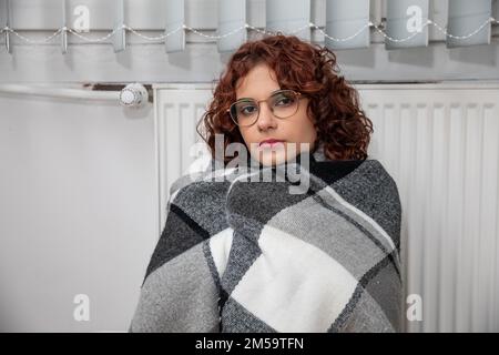 Una ragazza avvolta in una foulard spessa siede da un radiatore caldo. Riscaldamento centralizzato. Foto Stock