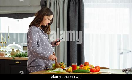 Donna incinta guarda la ricetta al telefono e prepara l'insalata Foto Stock