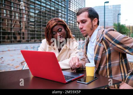 Due persone in viaggio d'affari lavorano insieme su un computer portatile mentre si siedono in un bar all'aperto. Foto Stock
