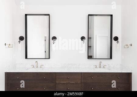 Un bagno di lusso con armadietto di legno scuro, ripiano in marmo e luci a lucernario intorno agli specchi quadrati. Foto Stock