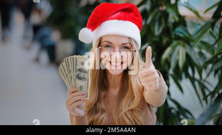 Felice ricco successo caucasica donna in rosso cappello di Natale di Santa e eyeglass in possesso di denaro contando denaro premio successo prima dello shopping Foto Stock