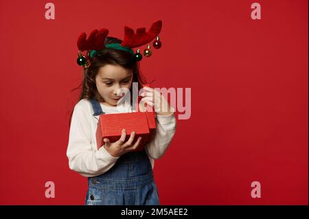 Sorpresa ragazza caucasica bambino indossando cervi corna cerchio, esprimendo la felicità mentre disimballaggio scatola regalo con regalo di Natale presente Foto Stock