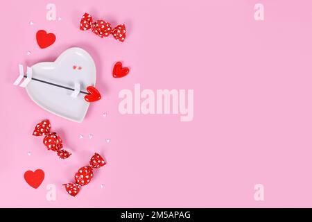 Caramella giorno di San Valentino carino, piatto a forma di cuore e frecce d'amore su sfondo rosa con spazio copia Foto Stock