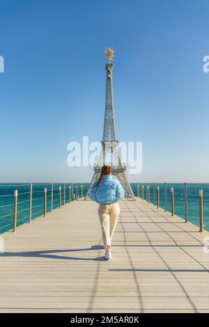 Grande modello della Torre Eiffel sulla spiaggia. Una donna cammina lungo il molo verso la torre, indossando una giacca blu e jeans bianchi. Foto Stock