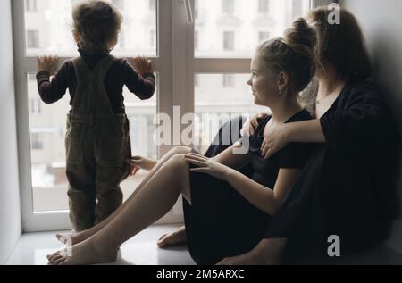 Mamma con bambini sul davanzale vicino alla finestra, guardando fuori per la strada. Mamma e sorella sono sedute sul davanzale, il ragazzo è in piedi Foto Stock