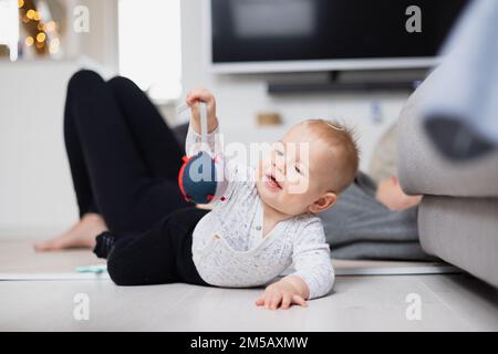 Momenti felici con la famiglia. Madre stesa comodamente sul tappeto dei bambini che guarda e sorveglia il suo bambino playinghis in salotto. Emozioni umane positive, sentimenti, gioia. Foto Stock